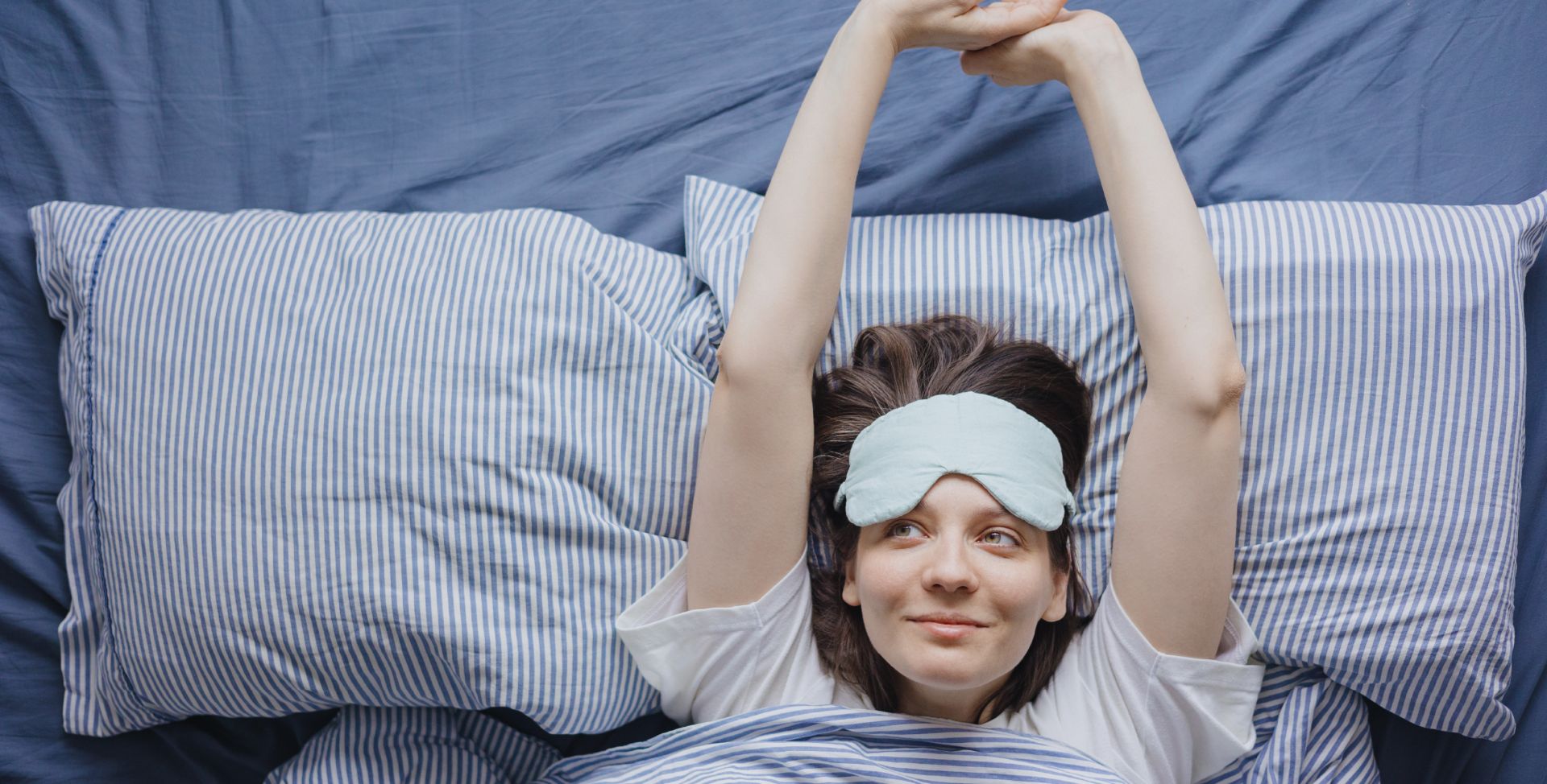 Eine Frau mit Schlafmaske streckt sich im Bett – bereit für gesunden Schlaf.
