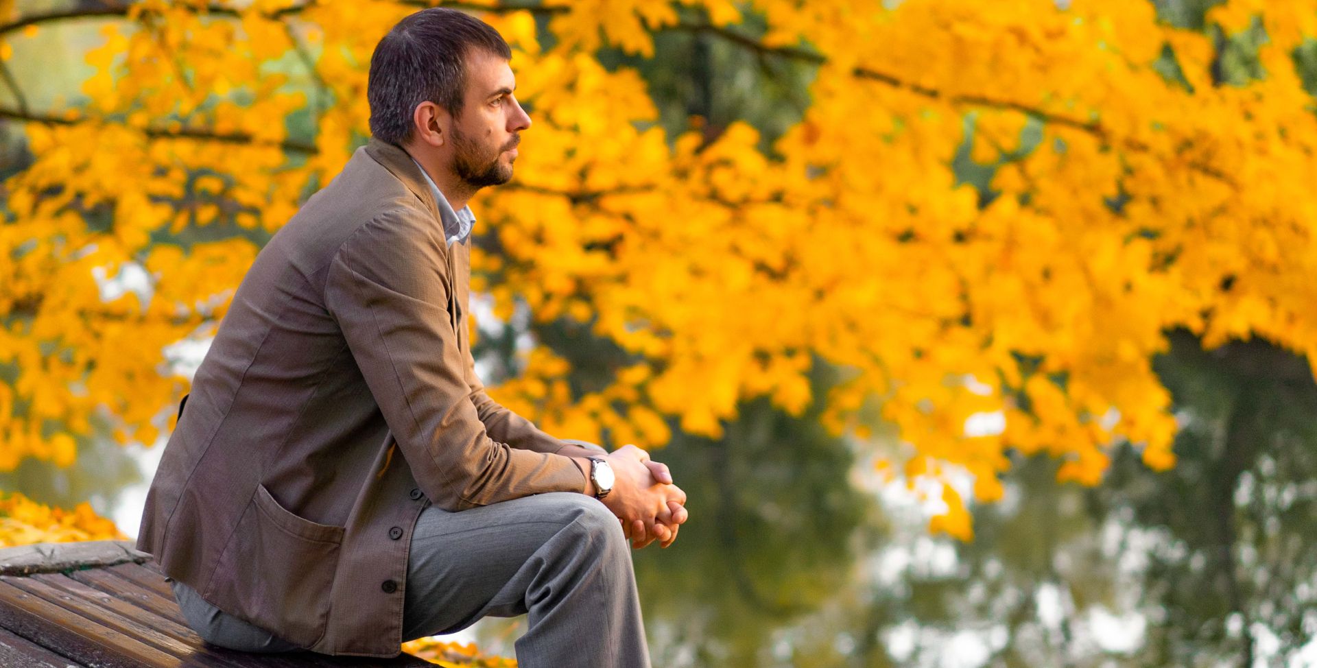 Ein Mann sitzt vor einem bunten Baum auf einer Bank und ist im Herbst- oder Winterblues.
