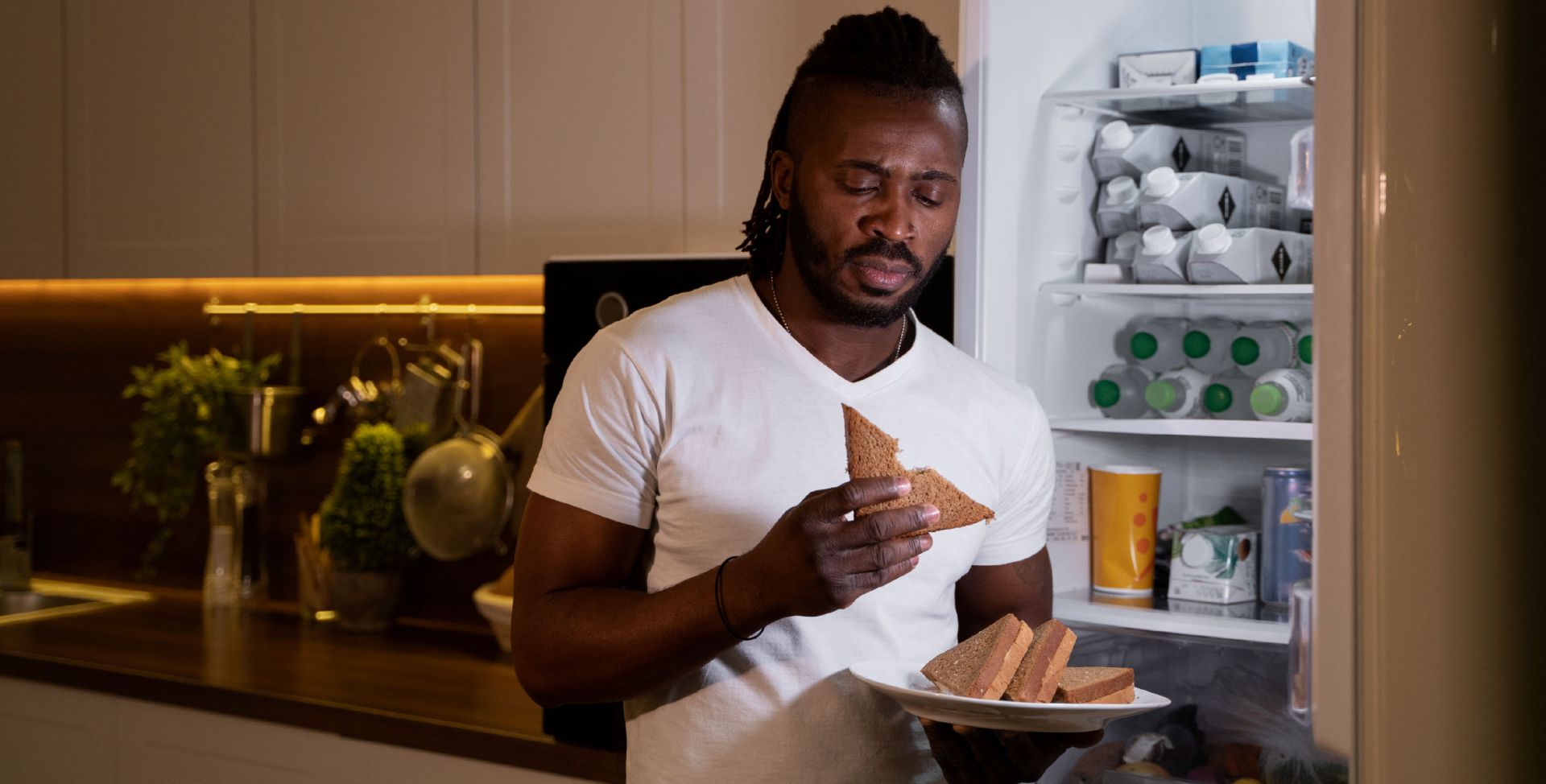 Ein Mann isst Sandwiches vor geöffnetem Kühlschrank. Sinnbild für die Hormone Ghrelin und Leptin.