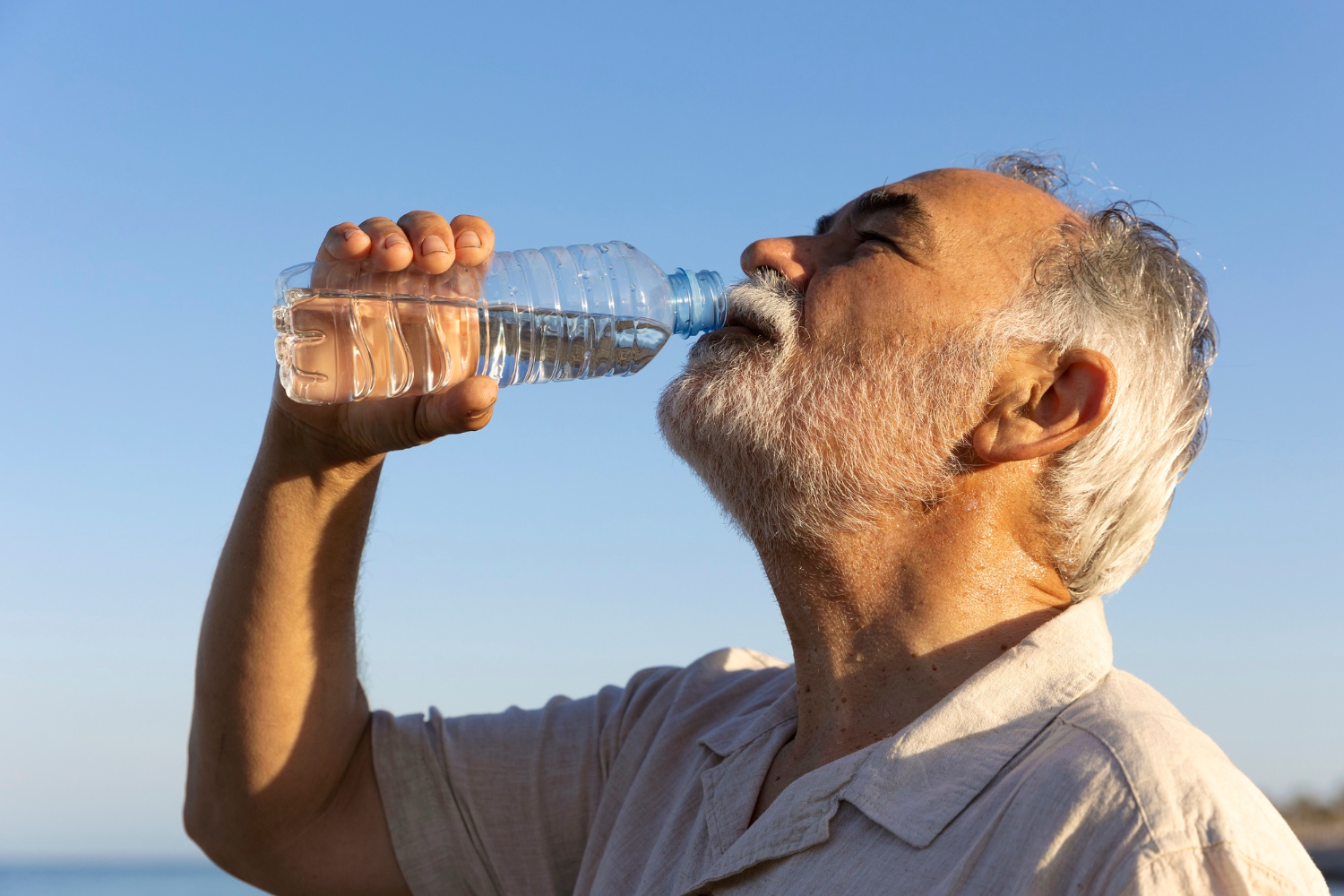 Ein älterer Mann trinkt in der Sonne Wasser aus einer Flasche, um Dehydrierung vorzubeugen.