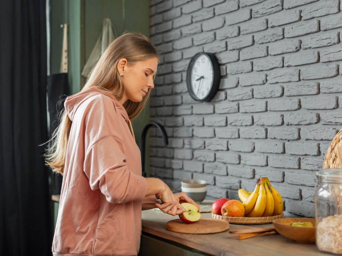 Eine Frau schneidet einen Apfel in der Küche vor einer Uhr. Sinnbild für Intervallfasten.